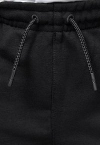 Pantaloni della tuta neri in tessuto testurizzato, dotati di chiusura con cordino e aglette nere, oltre a un cinturino elastico in vita. Design semplice e funzionale.