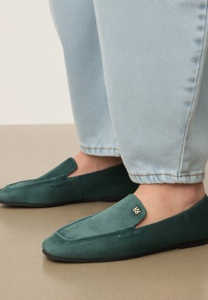 Mocassins en daim vert avec petit détail logo doré portés avec un jean bleu clair, vus de côté sur un sol beige.