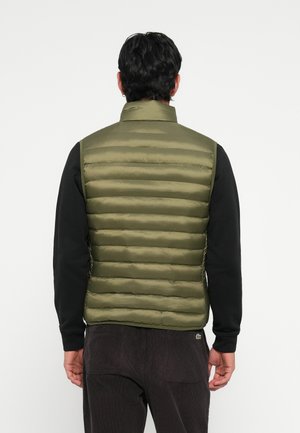 Olive green puffer vest with horizontal quilted design, high collar, and shiny texture, worn over a black long-sleeve shirt.