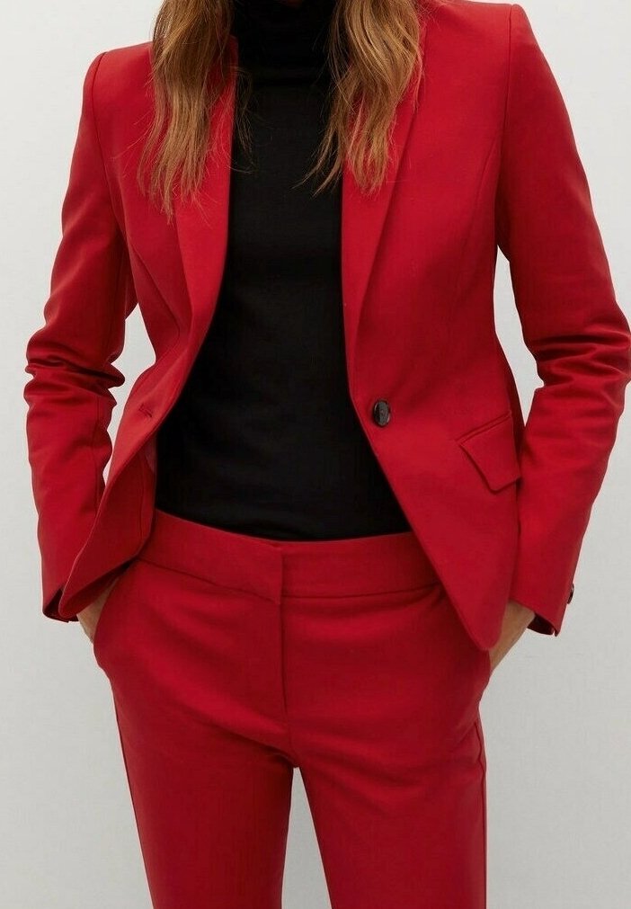 Femme portant un blazer rouge ajusté et un pantalon assorti avec un haut noir à col haut, les mains dans les poches, debout devant un fond uni.