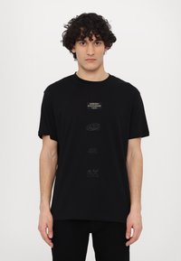 Juodas medvilnės marškinėlis su centriniu etiketu „ARMANI EXCHANGE 1991“ ir trimis įspausto logotipo dizainais žemiau, įvairių dydžių.