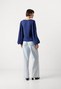 Blue textured top with puffed sleeves, paired with light blue wide-leg jeans and black block-heeled shoes. Simple, minimalist backdrop.