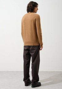 Beige gestrickter Pullover mit geripptem Muster, rundem Hals und langen Ärmeln, kombiniert mit dunkelbraunenweiten Hosen und schwarzen Schuhen.