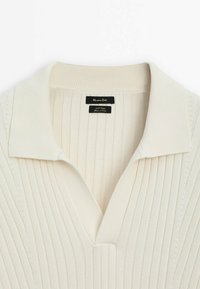 Chemise en maille côtelée couleur crème avec un large col et un décolleté en V, montrant l'étiquette de la marque cousue à l'intérieur du col.