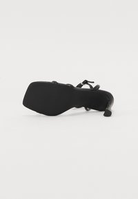 Scarpa nera con tacco alto caratterizzata da una parte superiore in pelle liscia, design con cinturini e suola in gomma testurizzata con tacco a punta.