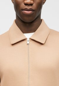 Εγγύς λήψη ενός ατόμου που φοράει μπεζ ζακέτα με φερμουάρ και γιακά, αποκαλύπτοντας εν μέρει ένα λευκό πουκάμισο από κάτω.