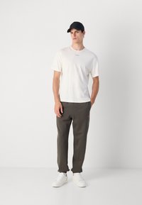Witte t-shirt met korte mouwen en logo, donkergrijze sweatpants met roze accent, en witte sneakers. Model draagt een zwarte pet.