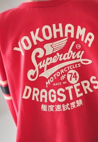 Rød bomuld T-shirt med stort, afskallet hvidt tekstdesign, der lyder "YOKOHAMA SUPERDRY MOTORCYCLES DRAGSTERS" med vinge- og racernummerdetaljer.
