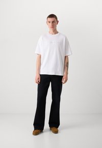 White cotton t-shirt with embossed "Calvin Klein" logo, paired with dark navy flared pants and brown suede shoes. Simple design and fit.