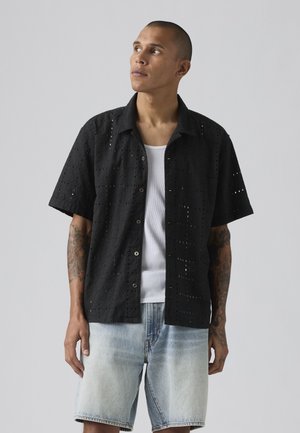 Mann mit Tattoos trägt ein schwarzes kurzärmliges Hemd mit Knopfleiste und Lochmuster, ein weißes Tanktop und helle Jeansshorts, blickt nach oben links.