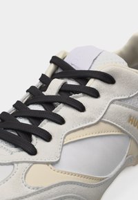 Gros plan sur une basket beige et blanche avec des lacets noirs, en suède et mesh, et un détail texte "HUE" sur le côté.