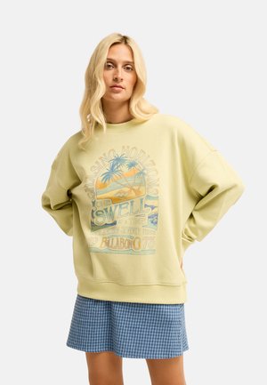 Blonde vrouw draagt lichtgele oversized trui met palmboom grafiek en tekst, gecombineerd met een blauw geruite rok, staand met handen achter.