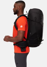 Svart ryggsäck med flera remmar och fickor, cylindrisk topp, fäst vid en person iförd en orange skjorta, sidoprofil vy.