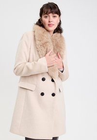 Manteau beige à double boutonnage avec un col en fausse fourrure, présentant des boutons noirs et des poches latérales, fabriqué à partir d'un matériau doux et texturé.