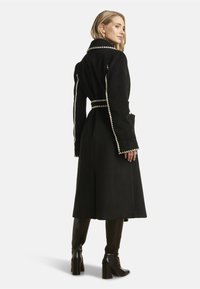 Long black wool coat featuring white stitching, a belted waist, and wide sleeves. The coat has a sleek, structured design with a split back.