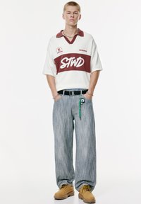 Kortærmet creme polo med maroon accenter og hvidt logo; løse lyseblå stribede jeans; brune arbejdstøvler; nøglering synlig på bæltet.