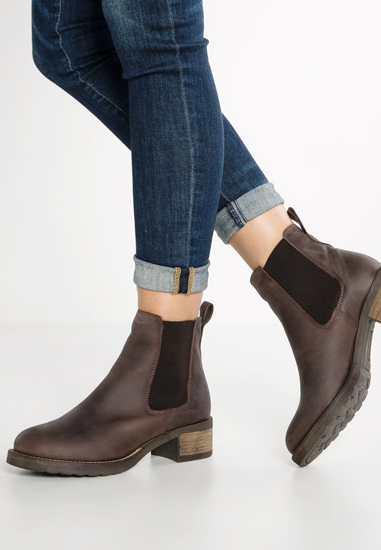 Stivali ankle in pelle marrone con lati elastici neri, indossati con jeans skinny blu scuro arrotolati su uno sfondo bianco.