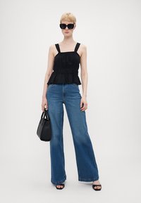 Svart peplum-top med brede stropper, sammen med høyt belte blå utsvingte jeans. Modellen holder en liten svart håndveske og har på seg store svarte solbriller.