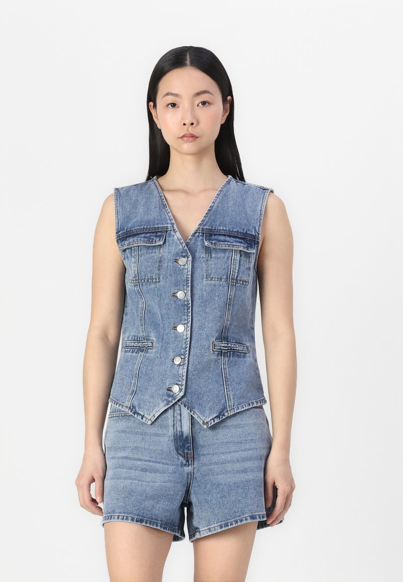 Lett blå dongerivest med V-hals, featuring to brystlommer, fem knapper og en tettsittende design. Kombinert med matchende dongerishorts.