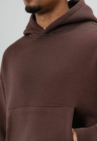 Bruine hoodie gemaakt van zachte stof, met een voorvak, geribbelde manchetten en een ontspannen pasvorm. De capuchon is bevestigd met een afgeronde halslijn.