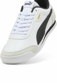 Sneaker en cuir blanc avec des accents noirs et gris, bout texturé et semelle plate en caoutchouc blanc. Équipé de lacets blancs et d'une languette de marque.
