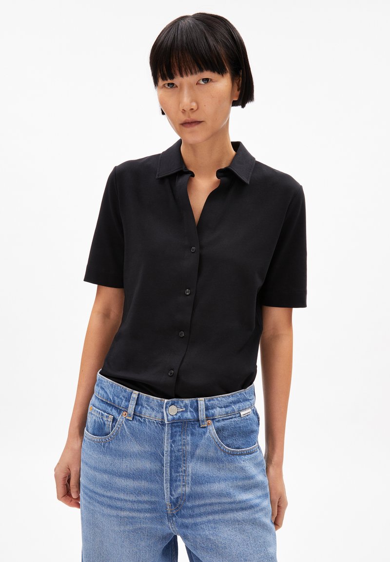 Black short-sleeve shirt with a collar, featuring a button front. Paired with light blue denim jeans, showing a classic fit and texture.