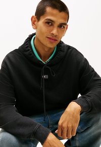 Felpa nera con cappuccio e cordoncino, con una piccola patch logo. Indossata sopra una camicia verde con colletto, abbinata a jeans di denim.