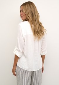 Bílá lněná košile s ohrnutými rukávy, s designem zadního yoku. Pruhované lehké kalhoty doplňují outfit.