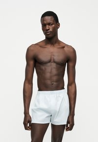 Άνδρας με σκούρο δέρμα χωρίς πουκάμισο, που φοράει λευκά σορτς με ελαστική μέση, στέκεται σε απλό φωτεινό φόντο, κοιτάζει μπροστά.