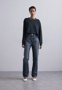Suéter verde oscuro con cuello redondo, combinado con pantalones vaqueros de tiro alto y corte holgado en azul. Presenta costuras blancas y zapatos de color crema.
