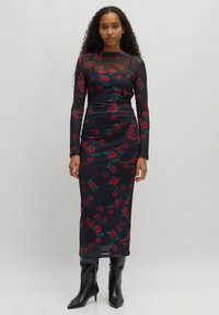 Langärmliges schwarzes Kleid mit rotem Rosenmuster, aus transparentem Material. Figurbetontes Design, knielange Länge, getragen mit schwarzen Stiefeletten mit Absatz.