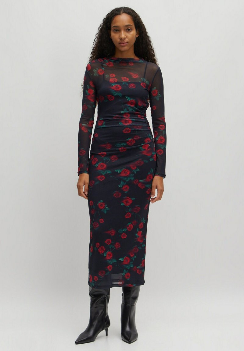 Langärmliges schwarzes Kleid mit rotem Rosenmuster, aus transparentem Material. Figurbetontes Design, knielange Länge, getragen mit schwarzen Stiefeletten mit Absatz.