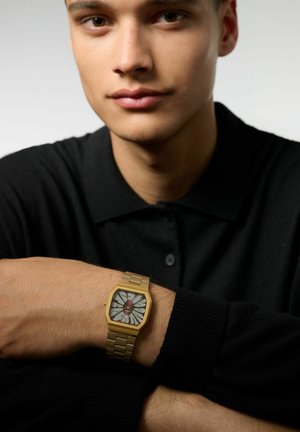 Montre analogique dorée avec un cadran géométrique, présentant des éléments de design contrastants en blanc et rouge, sur un bracelet en métal texturé. Polo noir.