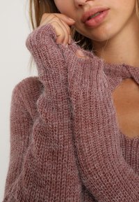 Jonge vrouw in een zachte mauve gebreide trui met lange mouwen, die een mouw dicht bij haar mond houdt en een gestructureerd patroon laat zien.