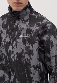 Giacca a motivi neri e grigi con colletto alto, chiusura con zip e un leggero marchio sul petto. Materiale leggero con una trama morbida.