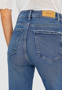 Rückenansicht einer Person, die hochgeschnittene blaue Jeans mit zwei Gesäßtaschen und einem beige Leder-Patch der Marke Stradivarius trägt.