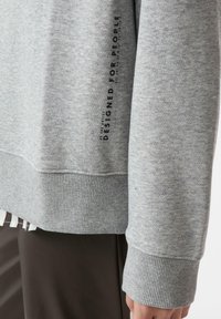 Jasnoszara bluza z prążkowanymi mankietami i ściągaczami, teksturowaną tkaniną oraz czarnym nadrukiem na rękawie z napisem "DESIGNED FOR PEOPLE."