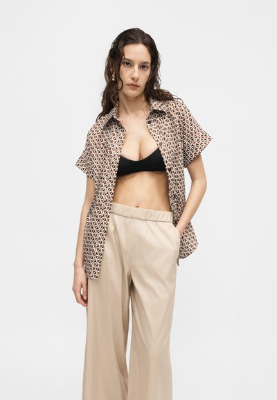 Γυναίκα με μακριά σγουρά μαλλιά που φοράει πουκάμισο με σχέδιο και κοντά μανίκια πάνω από ένα μαύρο κορμάκι και μπεζ φαρδιά παντελόνια, στέκεται με το ένα χέρι στην τσέπη.