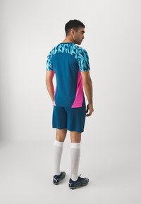 Sporttrikot mit blauem und türkisfarbenem geometrischen Muster, rosa Seitenpaneelen, passenden Shorts, weißen Socken und schwarzen Fußballschuhen mit grünen Akzenten.