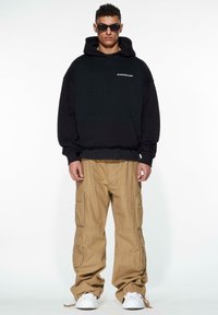 Sudadera negra de gran tamaño con bolsillo frontal y logo, combinada con pantalones cargo color beige que tienen múltiples bolsillos, llevada con zapatillas blancas.