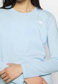 Vaaleansininen puuvillainen pitkähiha-paita, jossa pyöreä pääntie ja pieni valkoinen logo vasemmassa rintapäällisessä.