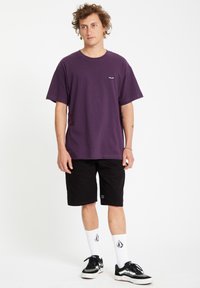 Paarse katoenen t-shirt met korte mouwen en een klein logo op de borst, gecombineerd met zwarte shorts, witte sokken en zwarte sneakers.
