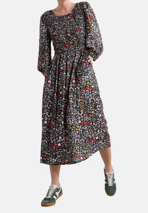 Femme portant une robe fleurie à manches longues, longue jusqu'aux chevilles, avec une taille froncée et des baskets vertes aux lacets et semelles blanches.