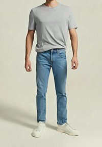 Homme portant un t-shirt gris clair à manches courtes, un jean slim bleu et des baskets blanches, debout devant un fond uni.