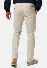 Chinos beige à coupe slim, dotés de deux poches arrière avec fermetures à bouton et d'une texture en coton lisse. Portés avec des espadrilles gris clair.