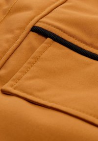 Bruine stof met een soepele textuur en zichtbare naden, met een zwarte ritssluiting en pocketdetail aan één kant.