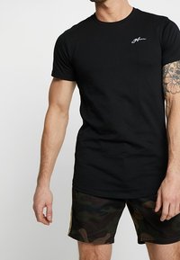 Man wearing black fitted t-shirt with small white logo on chest and dark green camouflage shorts with yellow stripe on side.