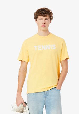 Gelbes Baumwoll-T-Shirt mit kurzen Ärmeln und rundem Ausschnitt, auf der Vorderseite mit fettem weißem Schriftzug "TENNIS". Kombiniert mit hellblauen Jeans.