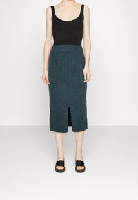 Dunkelgrüner, gerippter Midi-Rock mit einem Seitenschlitz, kombiniert mit einem schwarzen, eng anliegenden Tanktop und schwarzen Plateau-Sandalen. Schlichtes Design.
