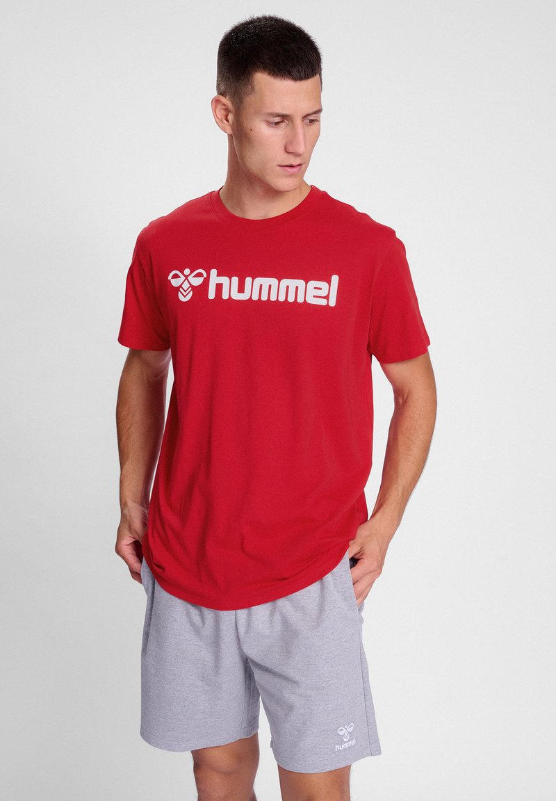 Rød t-shirt med korte ærmer og "hummel" logo i hvid, parret med lysegrå shorts. Glat stof, afslappet pasform, enkelt design.